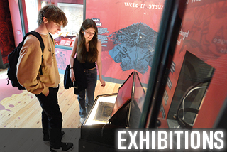 A young man and woman in their late teens examine an open, illuminated bible box with interest. They are in a museum gallery with red and blue walls and object cases.